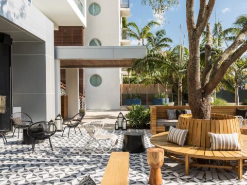 Modern outdoor seating area with geometric tiled floor, curved wooden bench, and black metal chairs surrounded by lush tropical plants and palm trees.