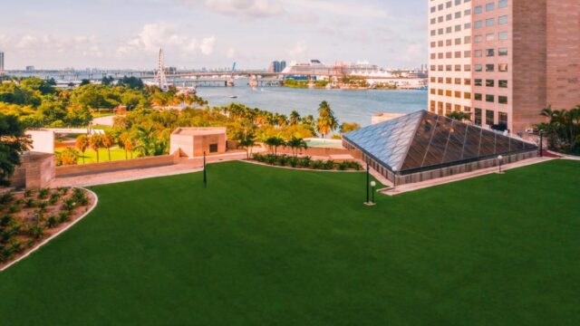 A lush green lawn slopes towards a modern glass pyramid structure, with a modern building and a cruise ship docked in the background under a bright sky.