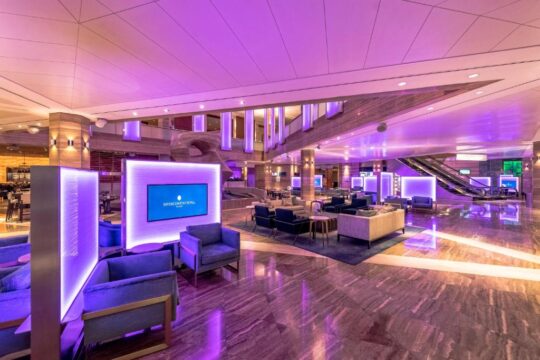 Luxurious hotel lobby with purple ambient lighting, modern seating areas, marble floors, and escalators.
