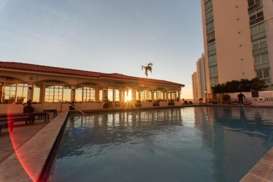 Sunrise over a resort pool and building, casting golden light on a restaurant with a red tile roof.
