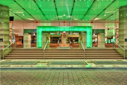 Modern hotel entrance with a grand staircase leading to glass doors, illuminated by vibrant green lights and framed by marble pillars.