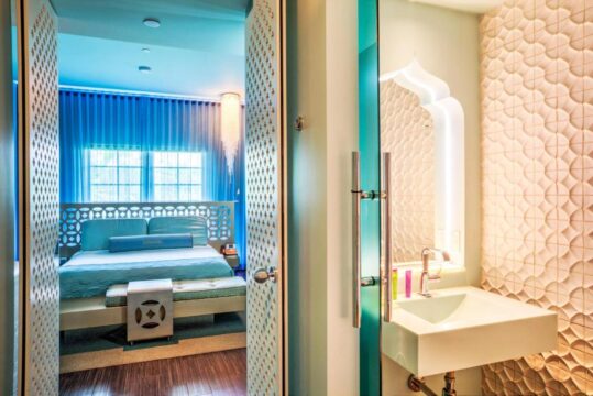 Luxurious hotel room with a vibrant blue color scheme and modern decor, featuring a king-size bed, elegant curtains, and a sleek bathroom with a white sink and textured wall.
