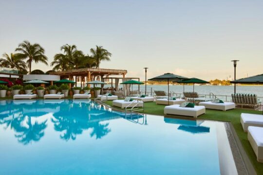 Luxury resort pool with white lounge beds and green striped umbrellas, overlooking a calm ocean under a clear sky. Palm trees and lush greenery frame the serene waterscape.