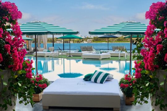 Luxury poolside loungers and striped umbrellas overlook a serene blue ocean, framed by vibrant pink bougainvillea. A plush daybed with green pillows sits in the foreground.