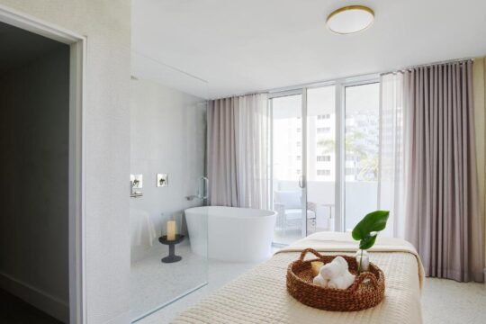 Luxurious spa room with a freestanding soaking tub, a massage table with rolled towels in a basket, and large glass doors opening to a balcony with seating.