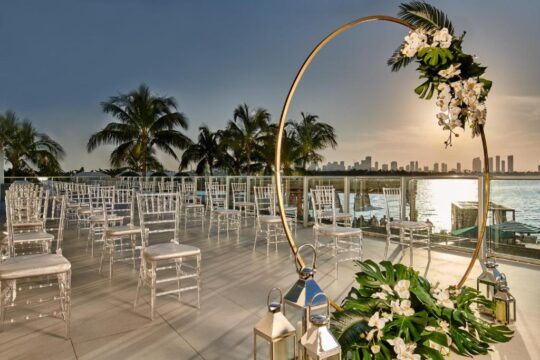 A romantic outdoor wedding ceremony setup with rows of clear chairs facing a golden circular arch adorned with white orchids and greenery, overlooking a serene ocean sunset.