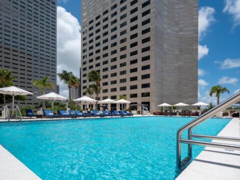 A vibrant blue swimming pool at a luxury hotel, surrounded by white lounge chairs and umbrellas, with modern skyscrapers in the background on a sunny day.