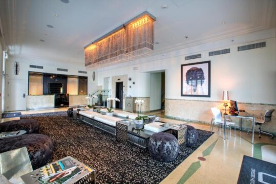 Luxurious hotel lobby with a large, ornate chandelier, modern seating areas with plush ottomans, and a striking framed artwork on the wall.