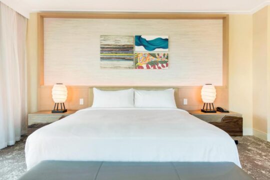 A luxurious hotel bedroom with a king-size bed, white linens, and modern bedside tables featuring lamps and artwork above the headboard.