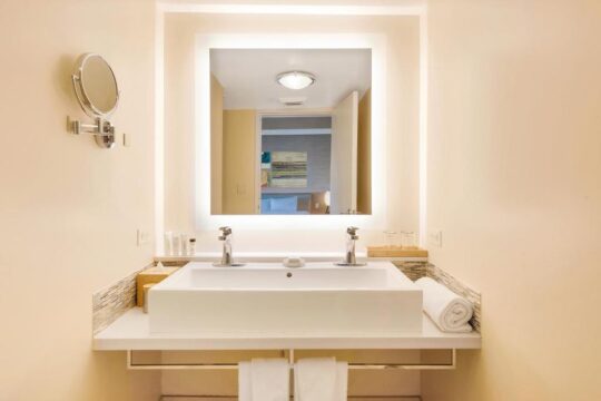 Brightly lit modern hotel bathroom vanity with a double sink, chrome faucets, and a large, illuminated mirror reflecting a hotel room. Amenities include rolled towels and toiletries.