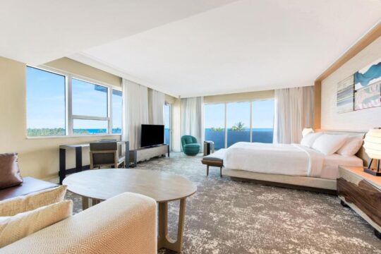 Spacious hotel suite with king-size bed, seating area, desk, and ocean view balcony. Modern decor, neutral tones, and large windows create a bright, luxurious atmosphere.
