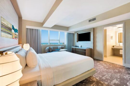 Luxurious hotel room with a king-size bed, ocean view window, and ensuite bathroom, featuring modern decor and plush carpeting.