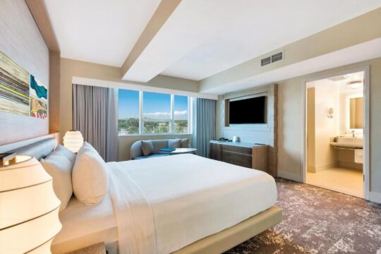 A luxurious hotel room with a king-size bed, large window overlooking a tropical landscape, and an open doorway to a modern bathroom.