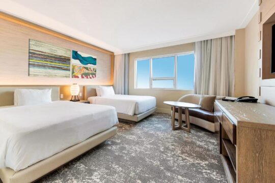 Modern hotel room with two queen beds, a large window with city views, and contemporary decor, featuring abstract art and a comfortable seating area.