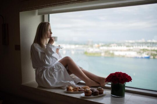 A blonde woman in a white robe enjoys coffee and pastries by a large window overlooking a city skyline and ocean, with a bouquet of red roses nearby.