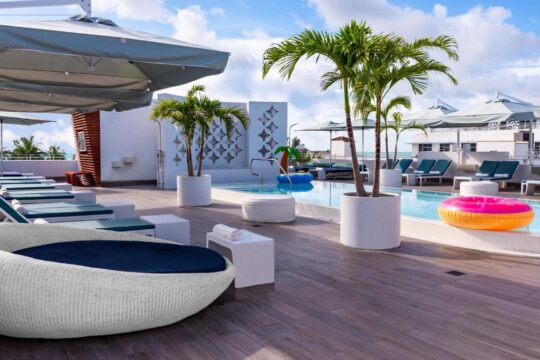 Luxury rooftop pool deck with white lounge chairs, palm trees, and inflatable pool toys, offering a vibrant and relaxing oasis with ocean views.