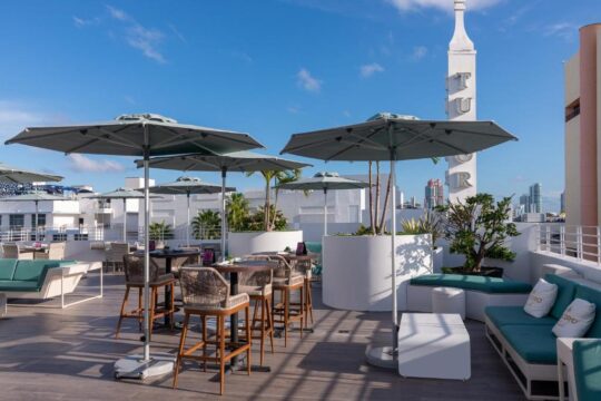 Rooftop bar with modern lounge seating, umbrellas, and tables overlooking a city skyline. White planters with greenery and a tall white tower with "TUR" visible complete the upscale outdoor dining ambiance.