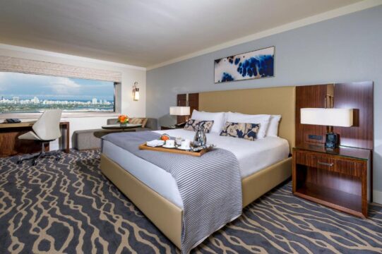 Luxurious hotel room featuring a king-size bed with a beige headboard, crisp white linens, and a striped throw blanket. A tray with coffee and snacks sits on the bed. A large window offers a city view, and a modern desk with a chair is visible.