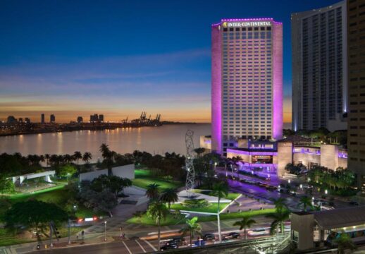 The InterContinental hotel in Miami glows with purple lights at dusk, overlooking Biscayne Bay with city skyline and port cranes in the background.