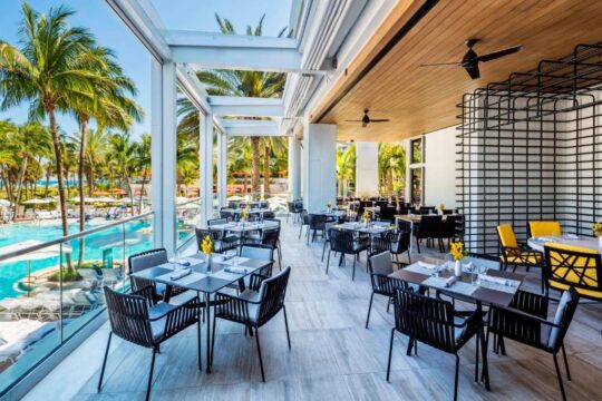 Elegant outdoor dining area at a tropical resort with palm trees, a pool, and modern seating, perfect for a luxury vacation experience.