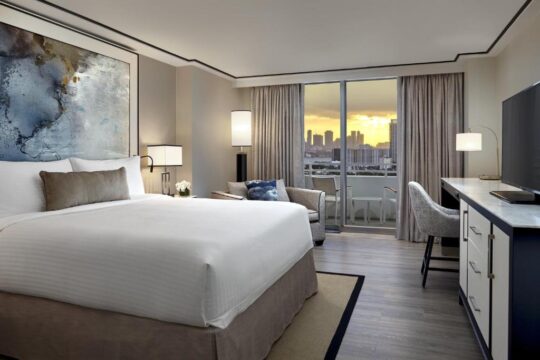 Luxurious hotel room with a king-size bed, modern desk, and a balcony overlooking a city skyline at sunset.