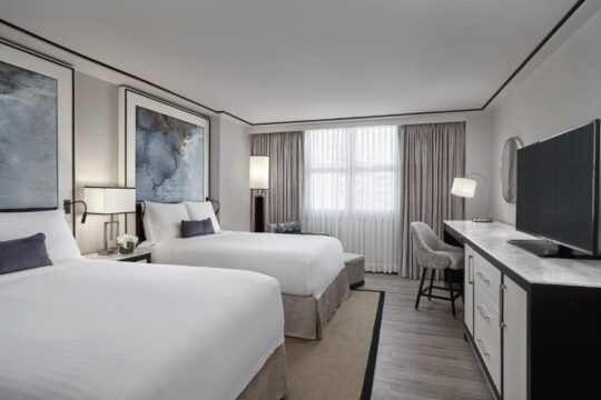 A hotel room with two queen beds, featuring modern decor, a large abstract artwork, and a desk with a TV.