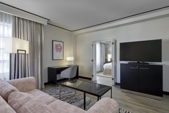 A modern hotel suite living area with a plush pink sofa, a black marble coffee table, and a desk with a chair. A large TV is mounted on a cabinet, and a doorway leads to a bedroom.