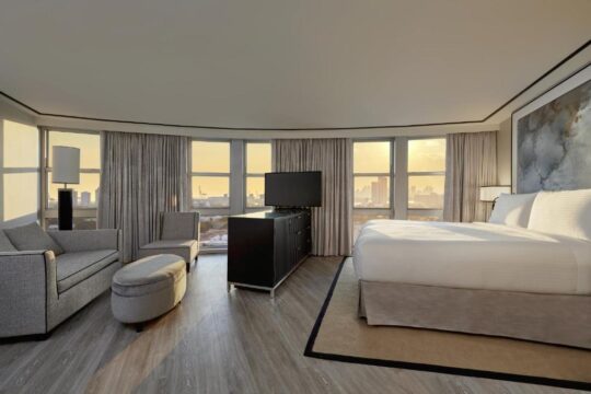 Luxurious hotel suite bedroom with a king-size bed, seating area, and panoramic city views through curved windows at sunset.
