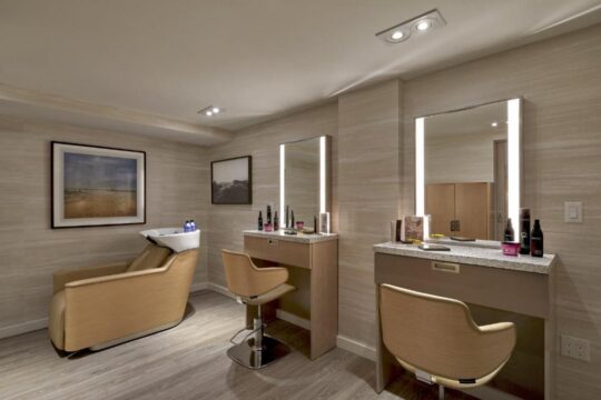 A modern salon interior featuring two styling stations with illuminated mirrors, comfortable chairs, and hair care products, alongside a shampoo station with a reclined chair.