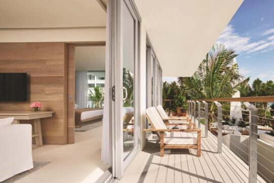Luxury hotel balcony with two wooden chairs and a railing overlooking palm trees and the ocean, with a modern bedroom visible through sliding glass doors.