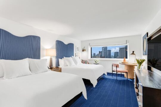 A hotel room with two queen beds, featuring blue wave-shaped headboards, a city view through a large window, and a bright blue carpet.