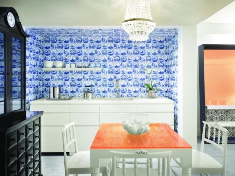 Modern kitchen with vibrant blue and white tiled backsplash featuring nautical scenes, white cabinetry, and an orange glass-top dining table with white chairs, illuminated by a crystal chandelier.