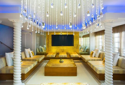Luxurious hotel lobby lounge with ornate crystal chandeliers, plush gold and cream sectional sofas, and a large gold mosaic wall behind a flat-screen TV.