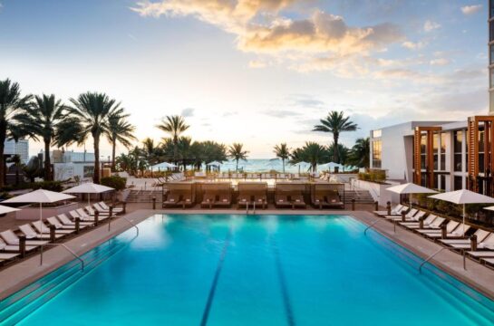 Luxury resort swimming pool at sunset, with palm trees, lounge chairs, and the ocean in the background.