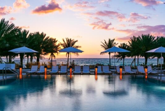 A serene infinity pool at sunset, lined with white lounge chairs and umbrellas, overlooks palm trees and a tranquil ocean with a colorful sky.