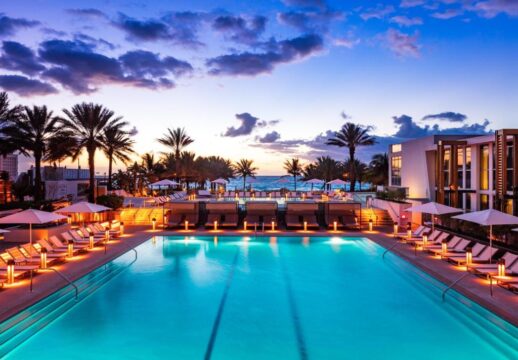 Luxury resort pool at sunset with palm trees and ocean view. Lounge chairs and umbrellas surround the illuminated turquoise water, creating a serene tropical ambiance.