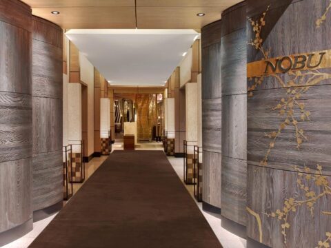 Entrance to Nobu restaurant with a long brown carpet runner leading down a hallway lined with textured wooden walls and decorative gold cherry blossoms. The Nobu logo is visible on the right wall.