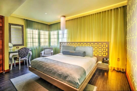 Luxurious hotel bedroom with a king-size bed, modern headboard, and vibrant yellow curtains. A vanity desk with a mirror and chair are visible.