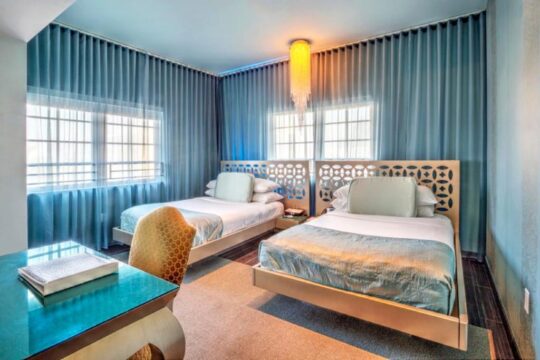 A modern hotel room featuring two queen beds with white linens and plush pillows, set against a backdrop of flowing blue drapes. A stylish desk with a teal surface and an ornate chair are visible in the foreground.