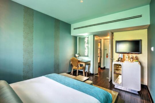 Modern hotel room with a king-size bed, turquoise walls, a desk with a chair, and a TV mounted above a mini-fridge.