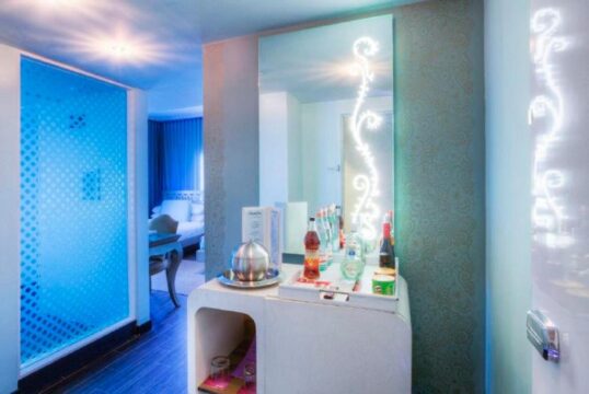 Modern hotel room with a mini-bar stocked with drinks, a large mirror with decorative lighting, and a frosted glass partition revealing a glimpse of a bed.