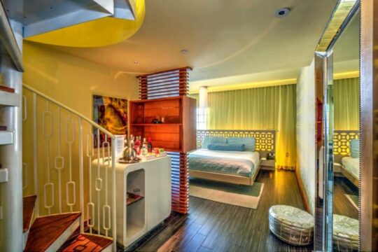 Luxury hotel suite with a spiral staircase, mini-bar, and a king-size bed with a teal headboard. Modern decor with yellow and green accents.