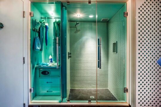 A modern hotel bathroom features a walk-in shower with textured green tiles and a glass door, adjacent to a compact dressing area with hanging clothes and a safe.