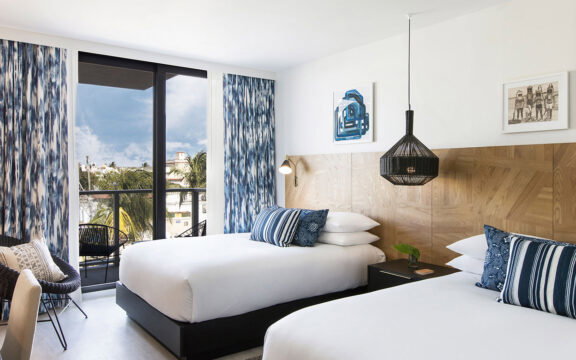 Bright hotel room with two queen beds, crisp white linens, and blue abstract patterned curtains. A wooden headboard and a large black pendant light add modern touches to the stylish decor.