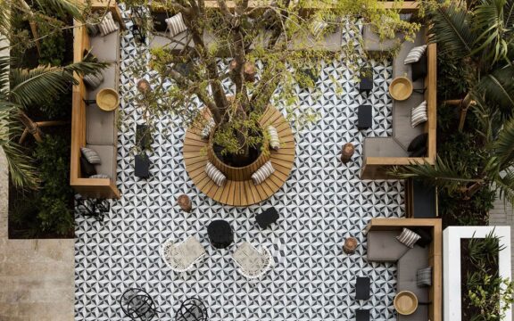 Aerial view of a modern outdoor lounge area with a large tree at its center, featuring geometric black and white tile flooring, wooden seating, and plush cushions.
