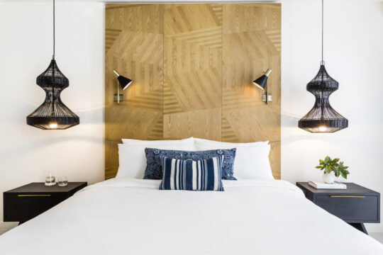 Luxurious bedroom featuring a king-size bed with crisp white linens, plush pillows, and a striking geometric wooden headboard. Two black woven pendant lights hang above black nightstands, flanking the bed.