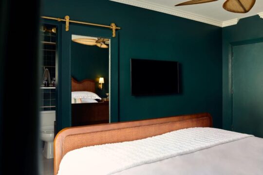 A luxurious hotel room features a king-size bed with a white duvet, a cane headboard, and a dark teal accent wall. A large flat-screen TV is mounted on the wall, and a sliding barn door with a mirror reflects a glimpse of the en-suite bathroom.