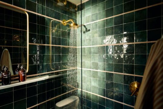 Luxurious emerald green tiled shower with brass fixtures, a rain showerhead spraying water, and a large mirror reflecting the opulent bathroom.