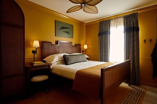 A cozy bedroom with a queen-size bed featuring a dark wood headboard and a burnt orange throw blanket. The walls are painted a warm mustard yellow, complementing the leopard print curtains and a ceiling fan with wooden blades.