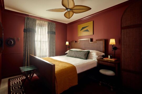 Luxurious hotel bedroom with a king-size bed, rich burgundy walls, a woven headboard, and a ceiling fan, creating a cozy and elegant atmosphere.